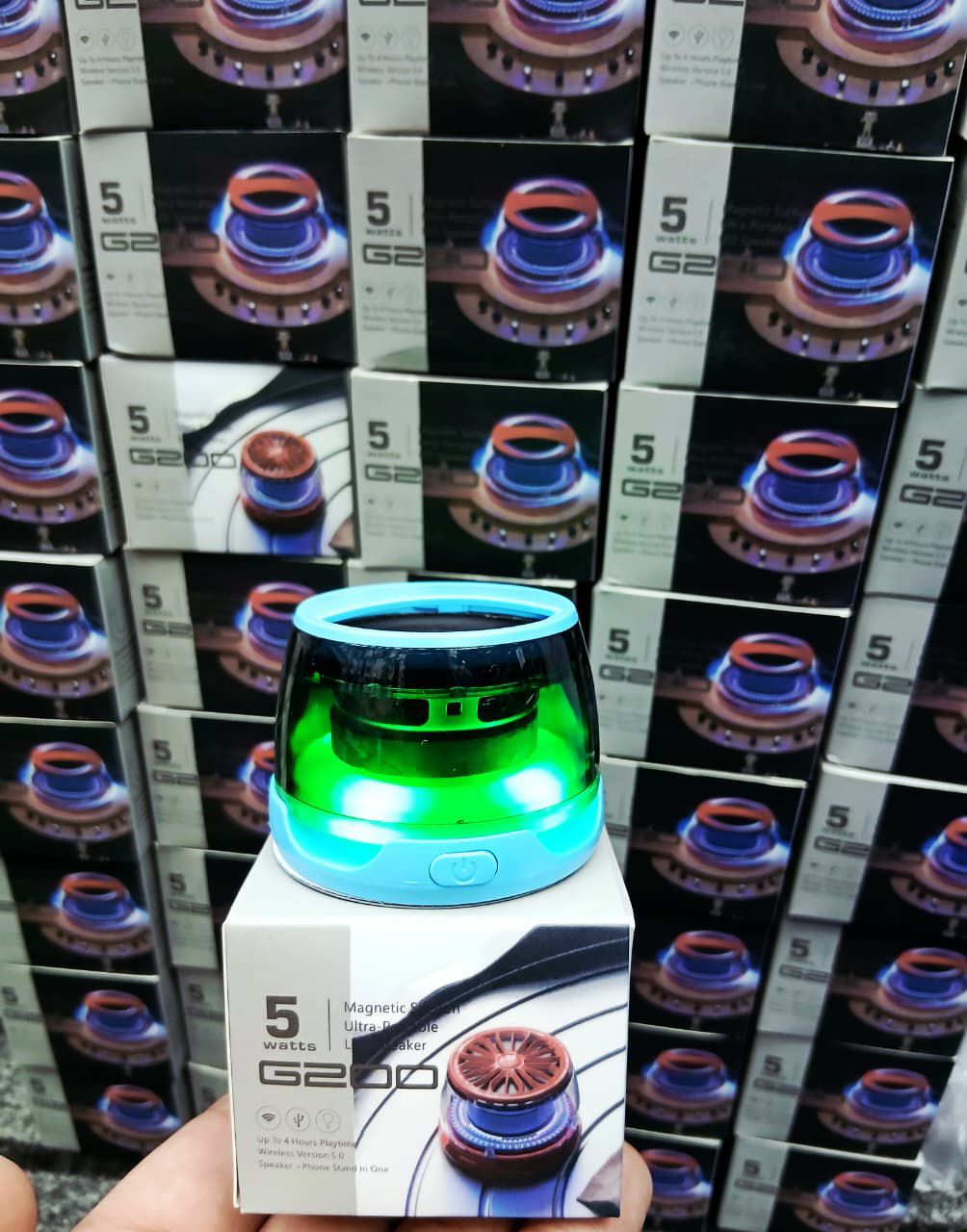Green and blue electronic device held in front of stacked boxes with similar devices.